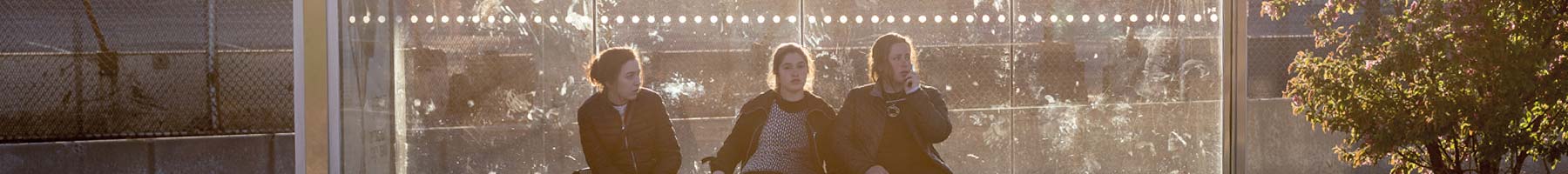 three girls sitting in a bus stop shelter in sunlight